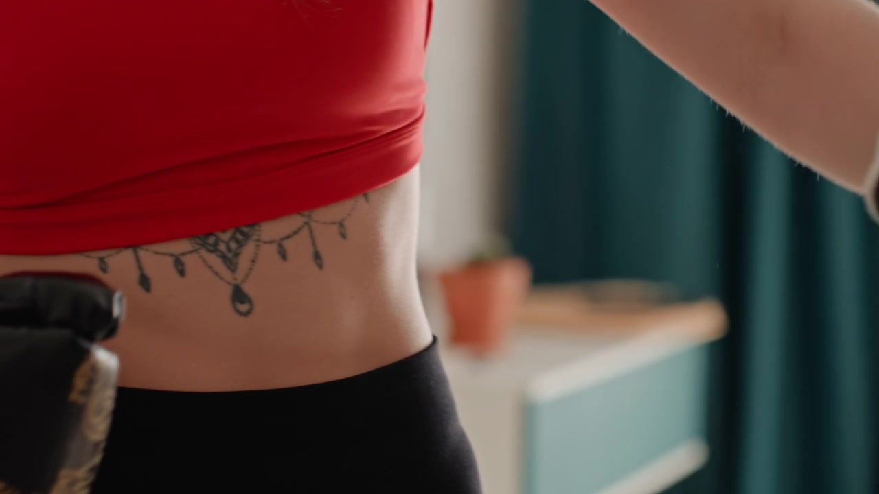White woman boxing glove closeups showing tattooed midriff, gold patterned gloves and strap adjustments, intimate preparation in bedroom, textured leather detail, focused hands tightening wrist