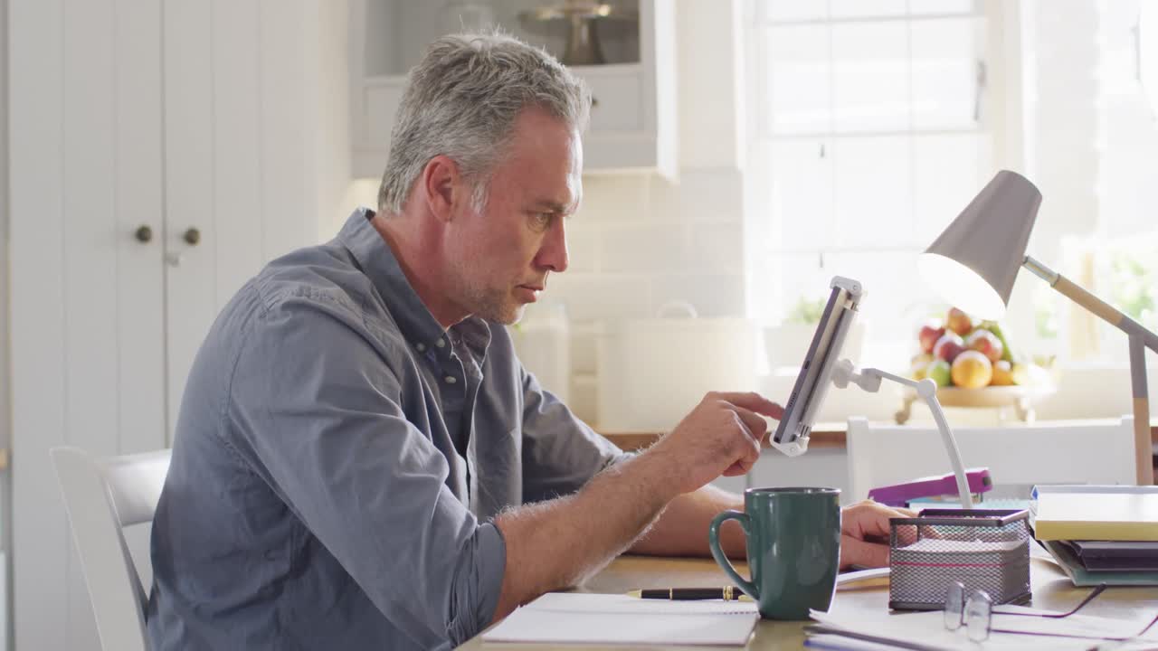 Happy caucasian man sitting at table in kitchen, using tablet