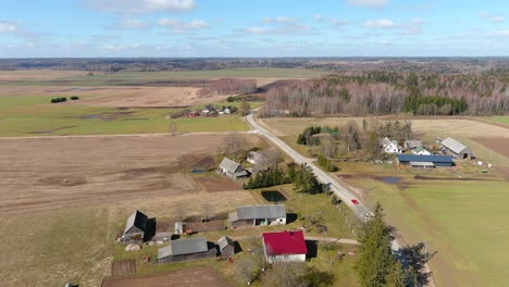 Aerial-View-of-Red-Car-Moving-on-Countryside-Road-Between-Farm-Buildings-and-Farmlands-in-Rural-Lithuania-on-Sunny-Day,-Drone-Shot