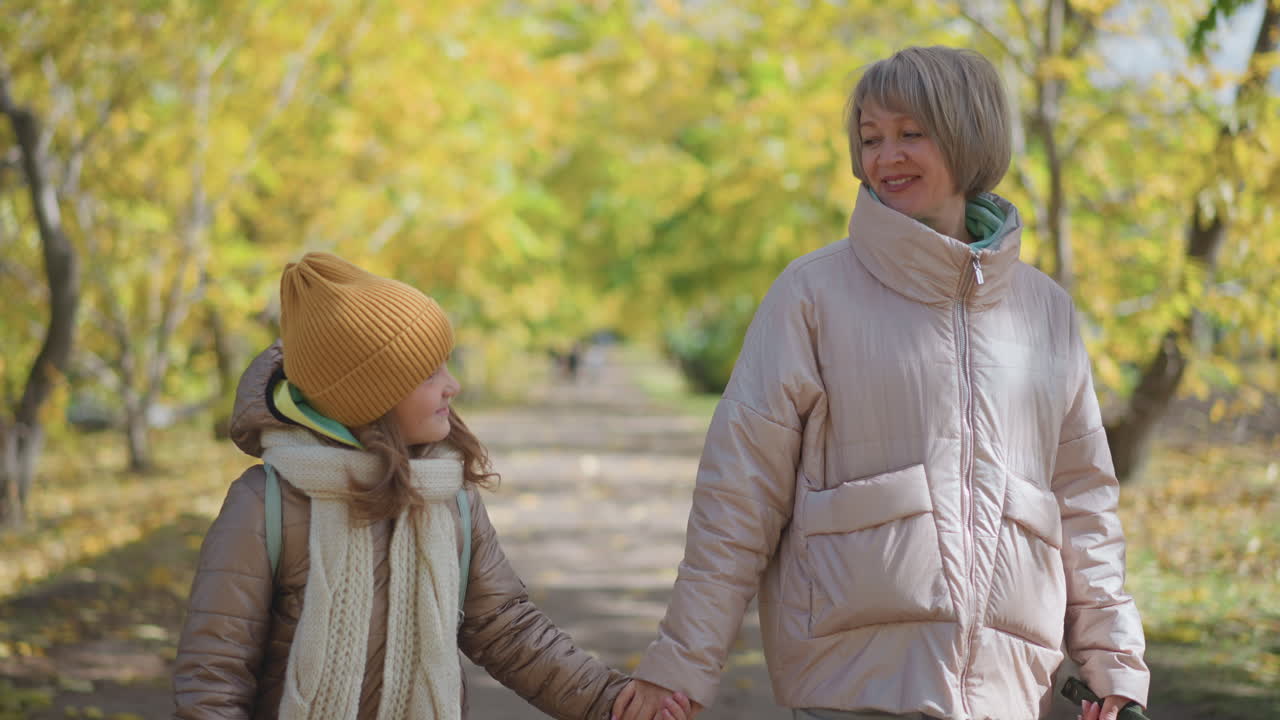 Mother lovingly interacts with son while holding hands and strolling together along peaceful autumn path surrounded by golden trees, sharing joyful expression