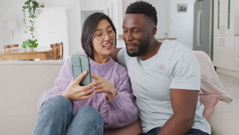 diverse couple sitting on couch and using smartphone in living room