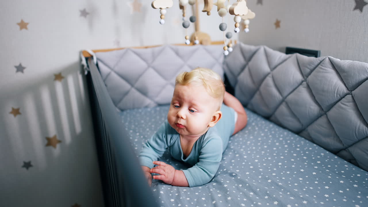 Little cute child lying on the belly. Adorable infant baby in bodysuit lies in cot under the crib mobile.