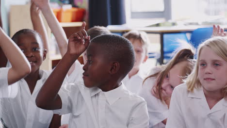 Group-Of-Elementary-Pupils-Wearing-Uniform-Sitting-On-Floor-Raise-Hands-To-Answer-Question-In-Class