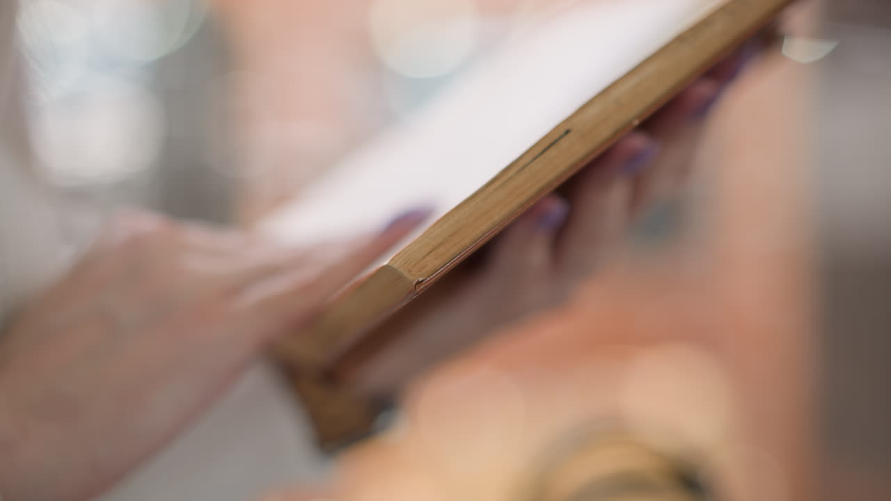close up of female hands with painted nails flipping worn pages of hardcover novel while seated on bench in busy mall, blurred modern retail interior creating calm focused reading moment