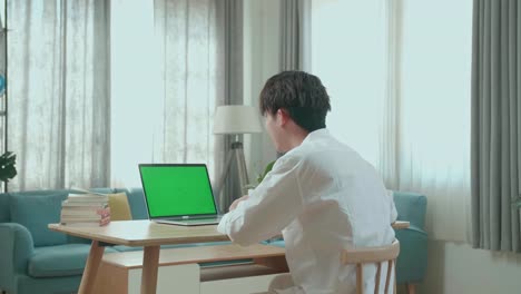 vista posterior de un estudiante asiático agitando la mano y hablando mientras usa una computadora de pantalla verde en casa