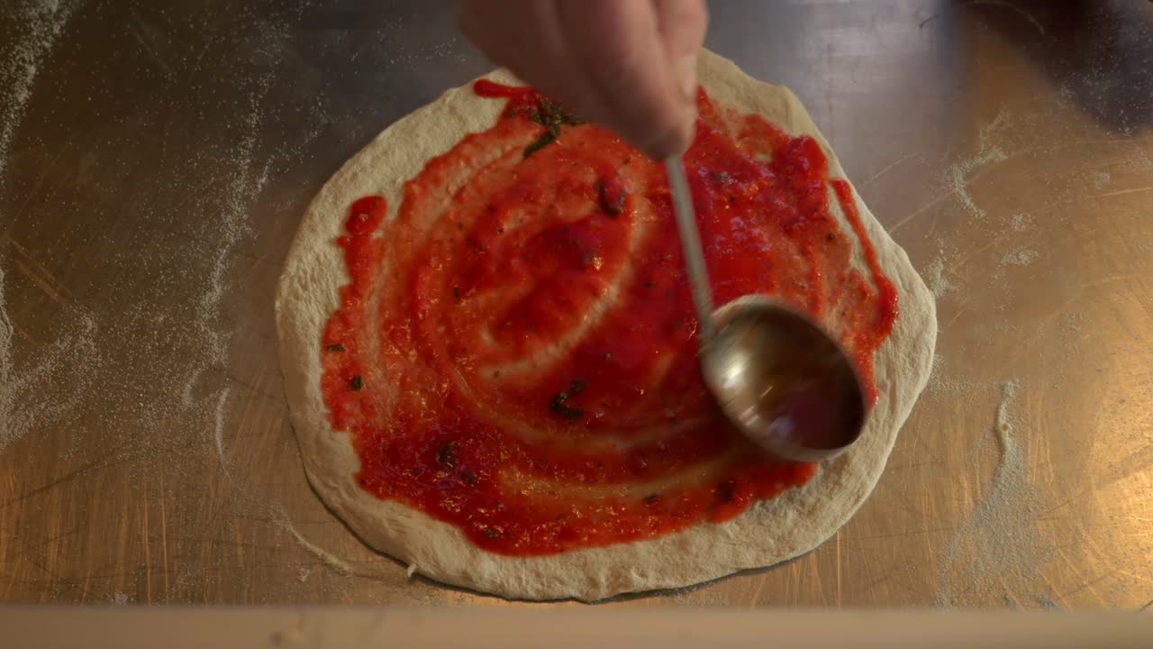 Inquadratura dall'alto di uno chef che aggiunge salsa per pizza all'impasto, in un'inquadratura luminosa e a mezzo primo piano.