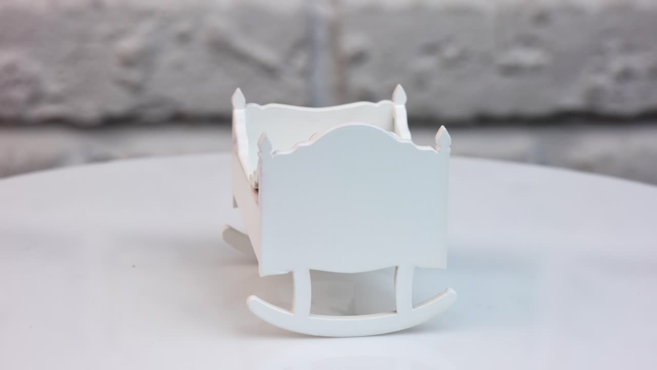 Tiny baby cradle at the rotating table. White toy crib with carved ornaments shaded pink. Close up. White wall background.