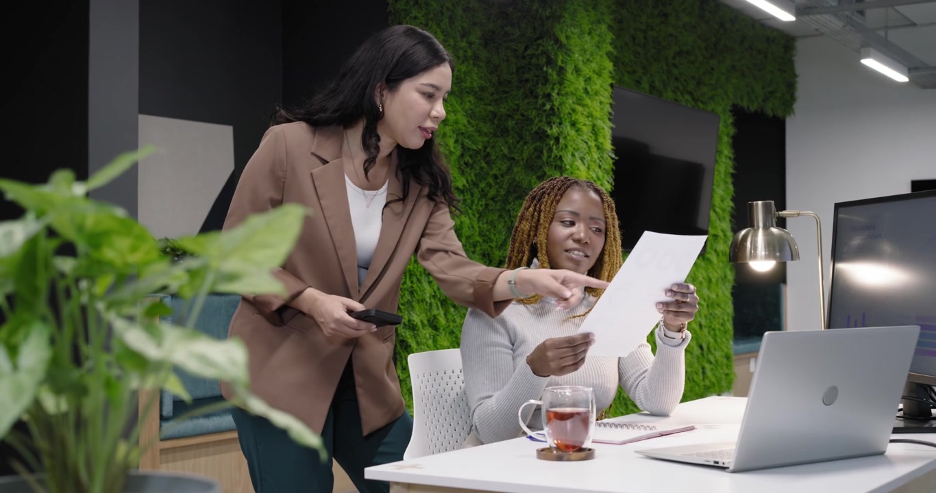 Female team at desk leaning, standing pointing paper with phone urging seated reading laptop