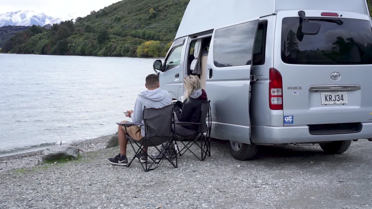 People camping by a lake with a camper van, enjoying breakfast in a scenic mountain setting.