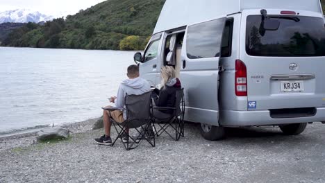 people camping by a lake with a camper van, enjoying breakfast in a scenic mountain setting.