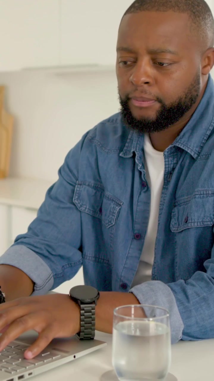 Vertical video: Studying laptop man in denim shirt leaning and typing at home kitchen, working