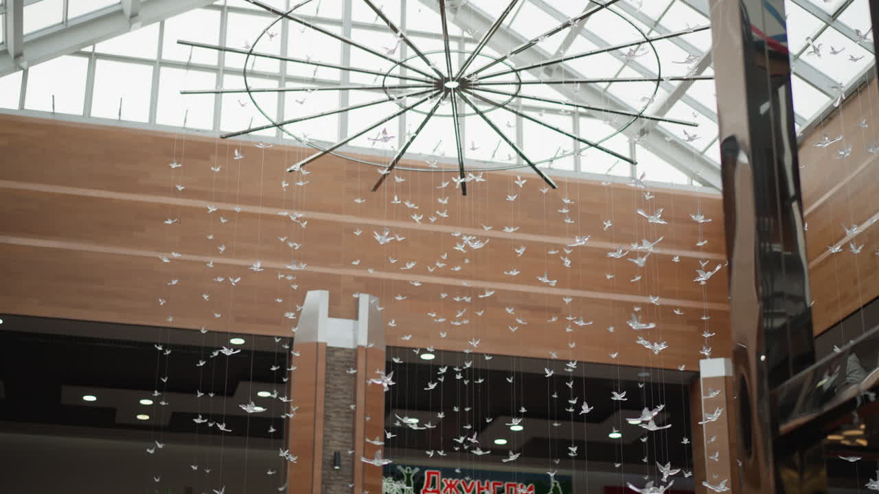 glass roof in modern mall adorned with suspended crystal sculptures fluttering like birds under skylight pouring light through steel beams onto wood paneled atrium creating elegant ambience
