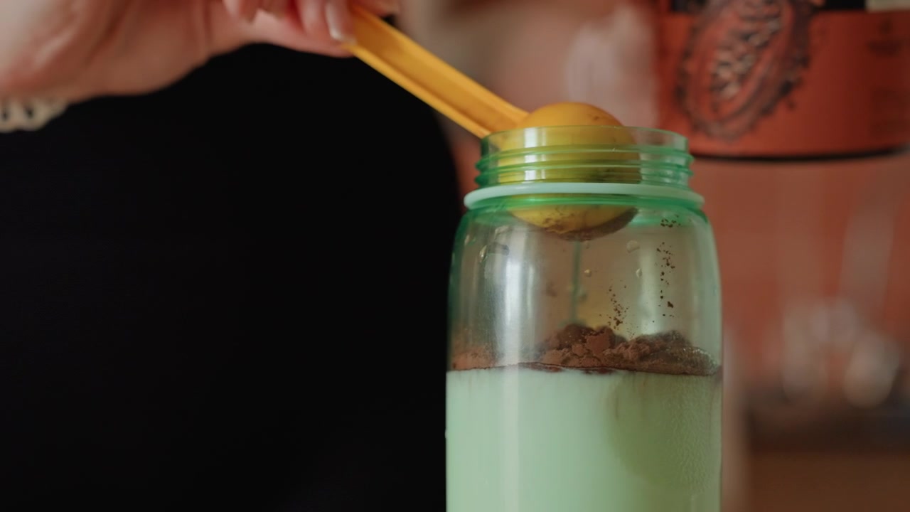 Donna che versa proteine in polvere in un barattolo nella cucina di casa, mani in primo piano che aggiungono polvere scura di integratore a liquido verde, top sportivo nero visibile, illuminazione calda e soffusa, rituale pre-allenamento calmo e concentrato