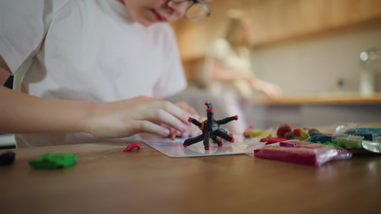 Boy in glasses shaping colorful clay at wooden table with handmade black clay sculpture in foreground, mother blurred in background, symbolizing creativity, imagination, concentration artistic indoor activity