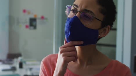mujer caucásica pensativa que usa una máscara facial con la mano en la barbilla en una oficina moderna