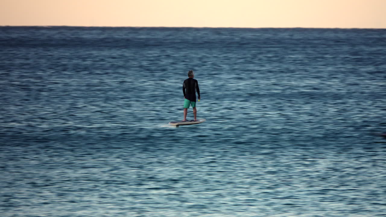 Middle age man in green shorts riding an electric foil board on the ocean at sunset.