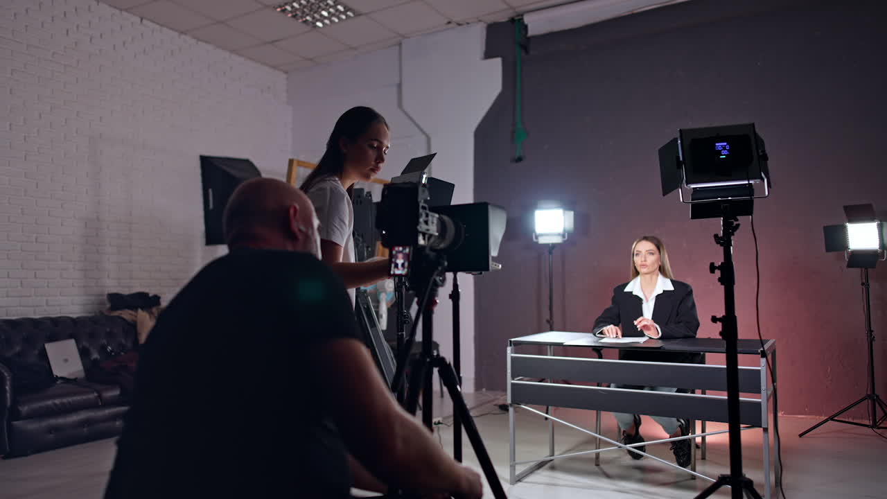 Male photographer sitting behind the camera and giving a sign to a lady sitting at desk. Studio footage of a program. Backstage work.