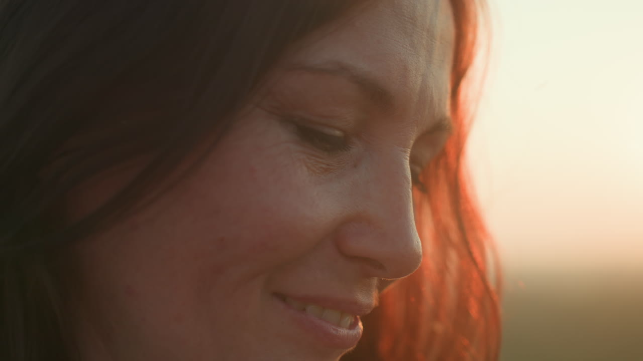Close up of young woman smiling after taking photo outdoors during golden hour with sunlight highlighting hair and soft breeze in serene natural setting, expressing joy