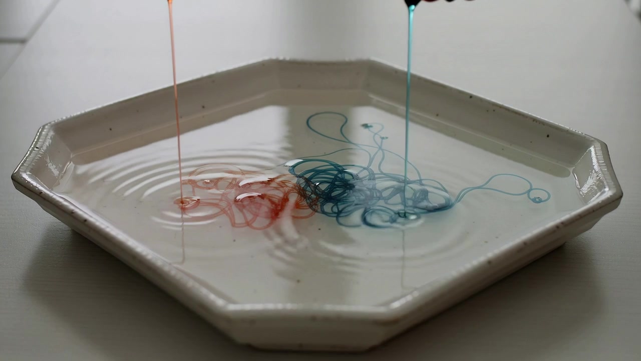 Dripping dropper sending orange-teal streams into white hex plate with water on tabletop for demo