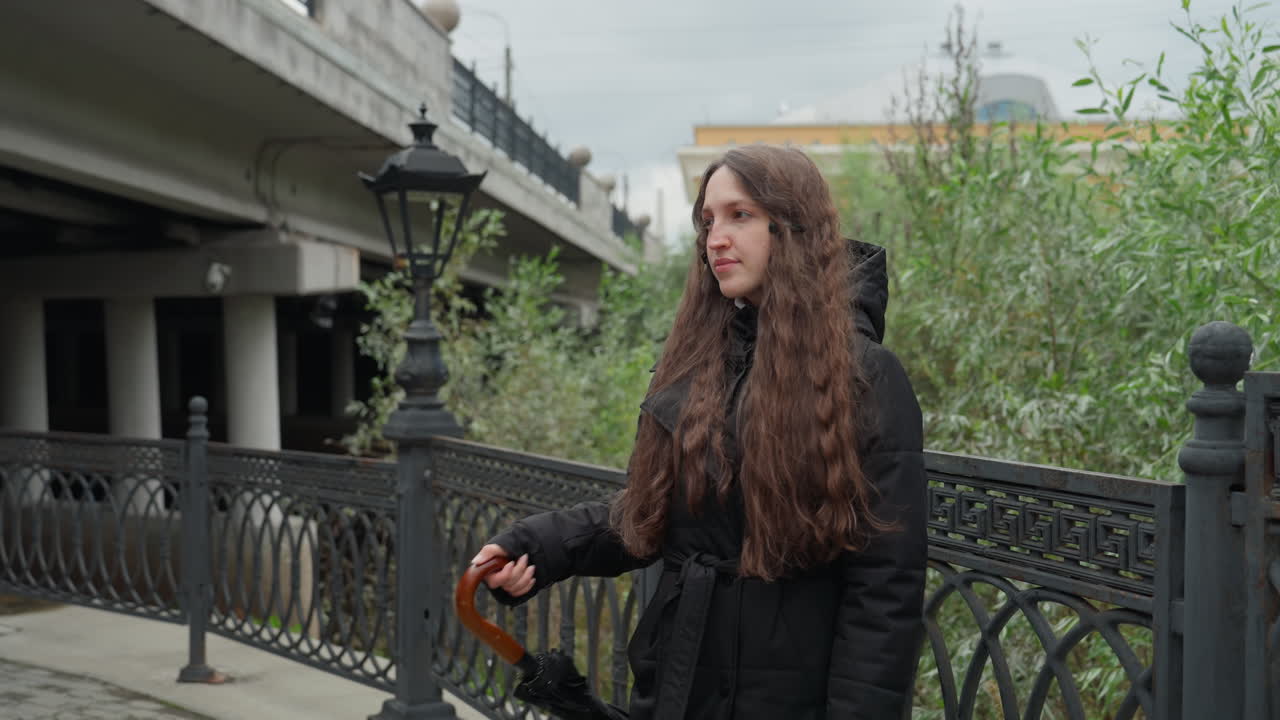 Giovane donna che tiene un ombrello su un ponte, passerella cittadina con ringhiera in ferro e lampioni, cielo nuvoloso, lunghi capelli ricci che fuoriescono dal cappuccio del parka nero, un dolce sorriso durante la passeggiata, fogliame autunnale