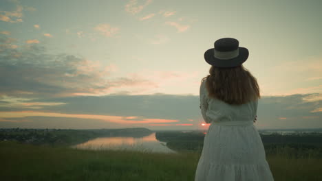 close-up back view of a woman in a white dress and hat, arms folded, standing alone in a serene grassy field, gazing out over a tranquil lake during sunset, under a soft, pastel-colored sky