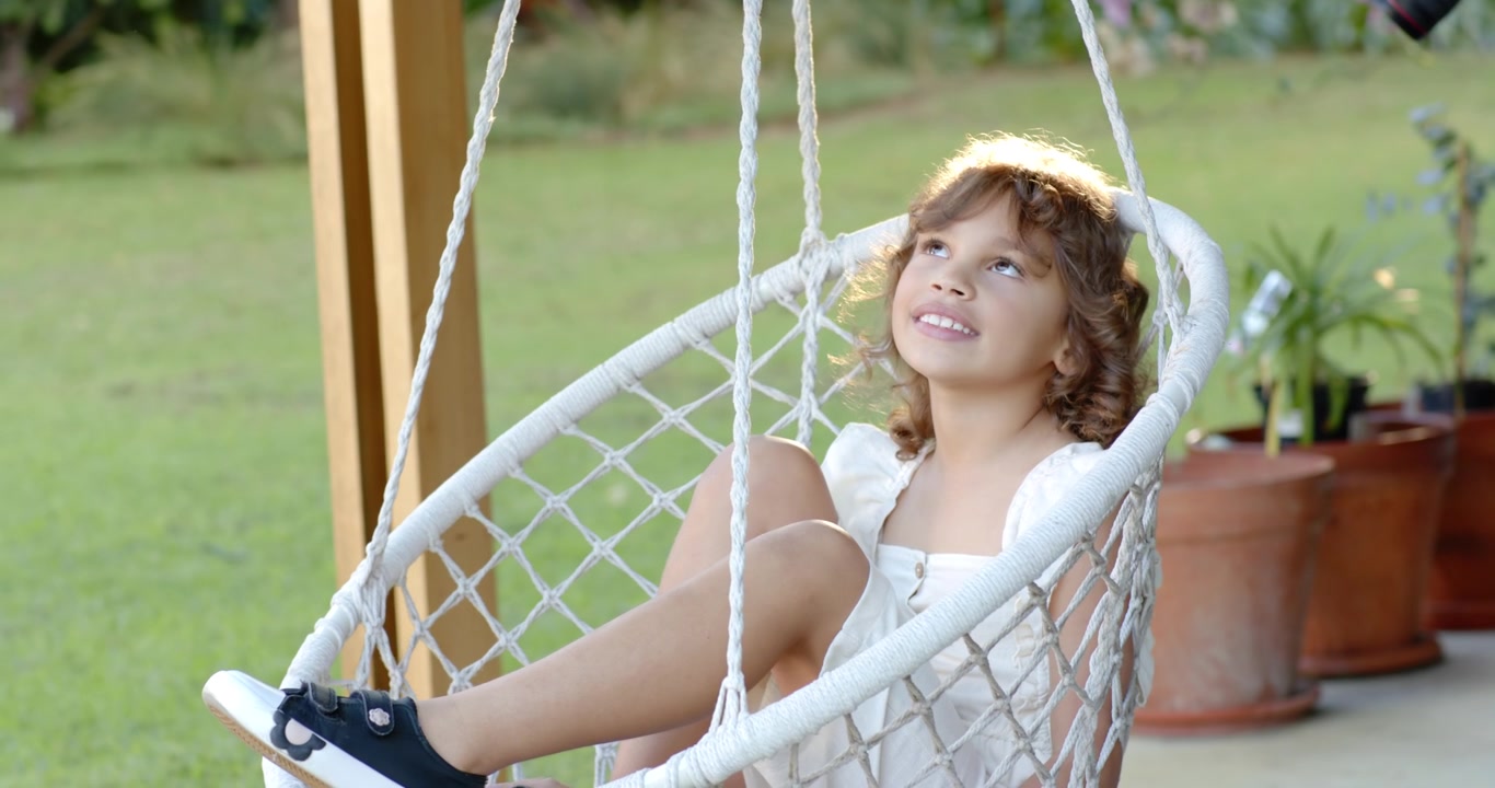 Bambina afroamericana di 6 anni che si dondola su una sedia sospesa intrecciata sul portico, seguendo la luce del sole.