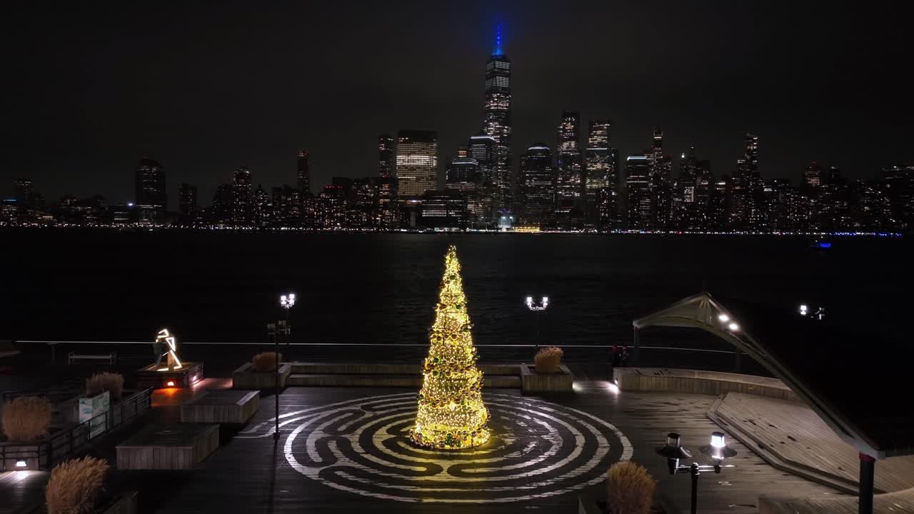Illuminated Christmas tree on Hoboken Pier overlooking Manhattan skyline. One World Trade Center and Hudson River backdrop at night. Stunning aerial of holiday ambiance.