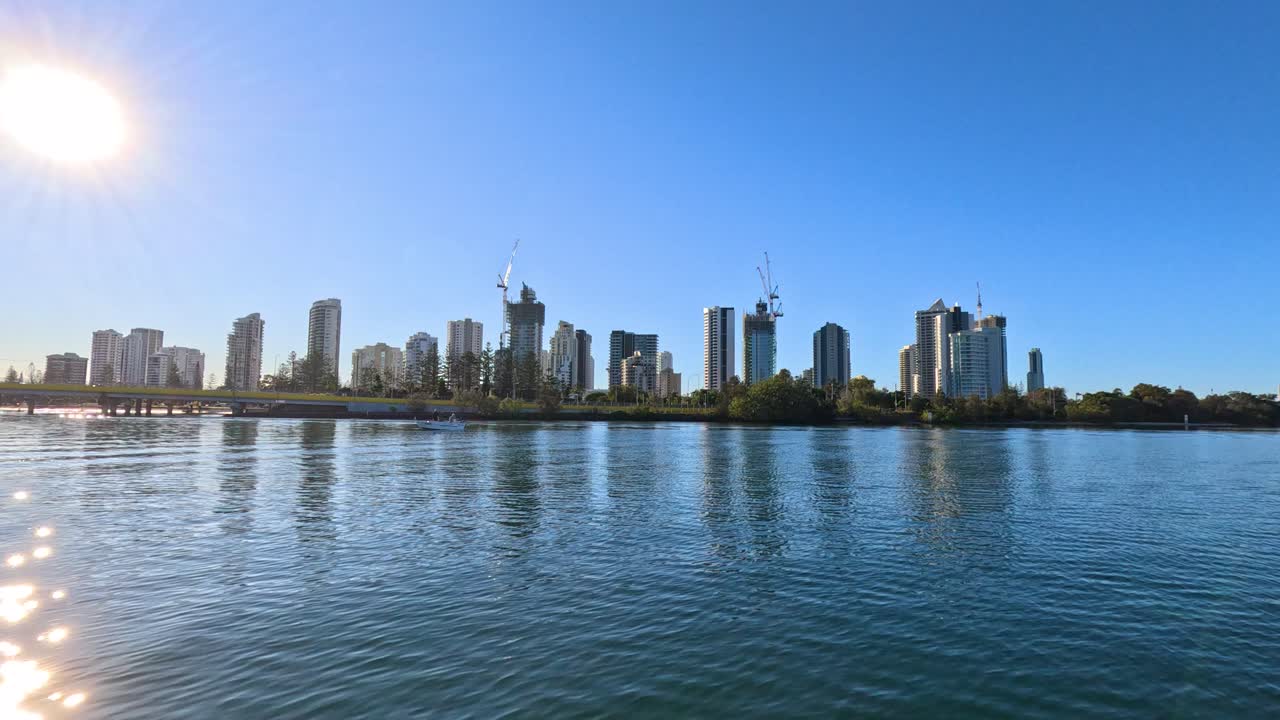 A camera glides smoothly over calm river water toward a speed limit sign, revealing Gold Coast city skyline under bright, clear daylight