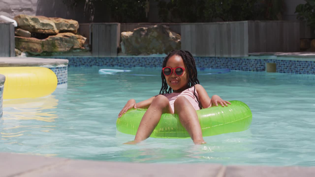 feliz niña afroamericana con gafas de sol flotando en un anillo inflable en la piscina
