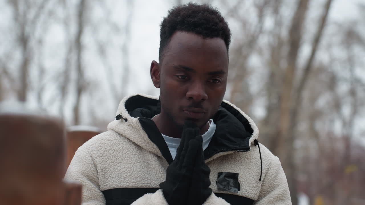 Young boy wearing black gloves and white winter jacket outdoors in cold weather, hands pressed together in deep thought, with a blurred background of bare trees and snow