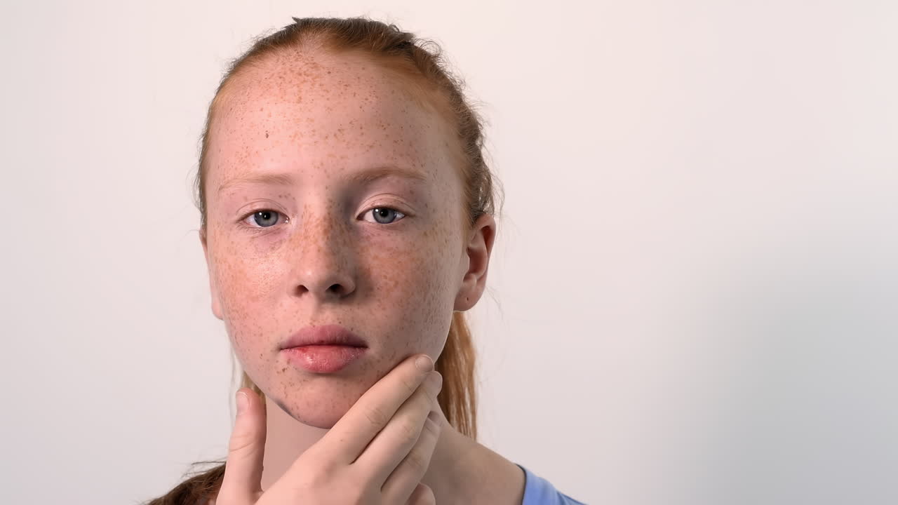 Red haired girl with blue eyes touching face full of freckles. Looking into the camera. White background. Tighten hairs