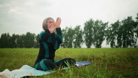 woman seated on yoga mat in misty field practicing yoga with eyes closed, hand twisting, and thumb touching forehead, the backdrop includes a cloudy sky with trees lined up along the horizon