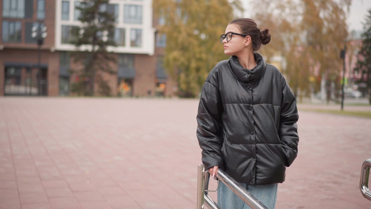 Young woman with glasses stands outdoors in public square wearing black puffer jacket and blue jeans. Woman holds onto metal railing while looking around urban environment which features
