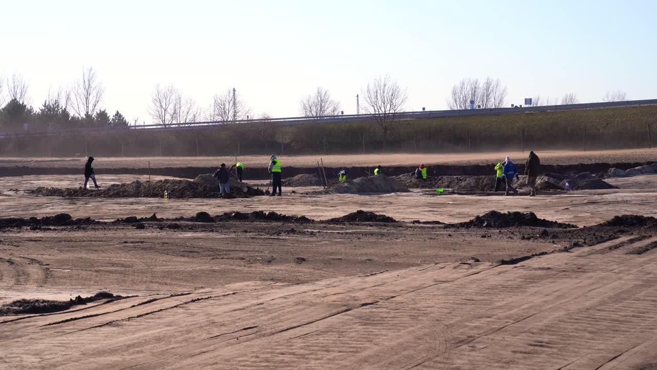 Dust blows across workers digging at archaeology site. Wide angle. Establishing