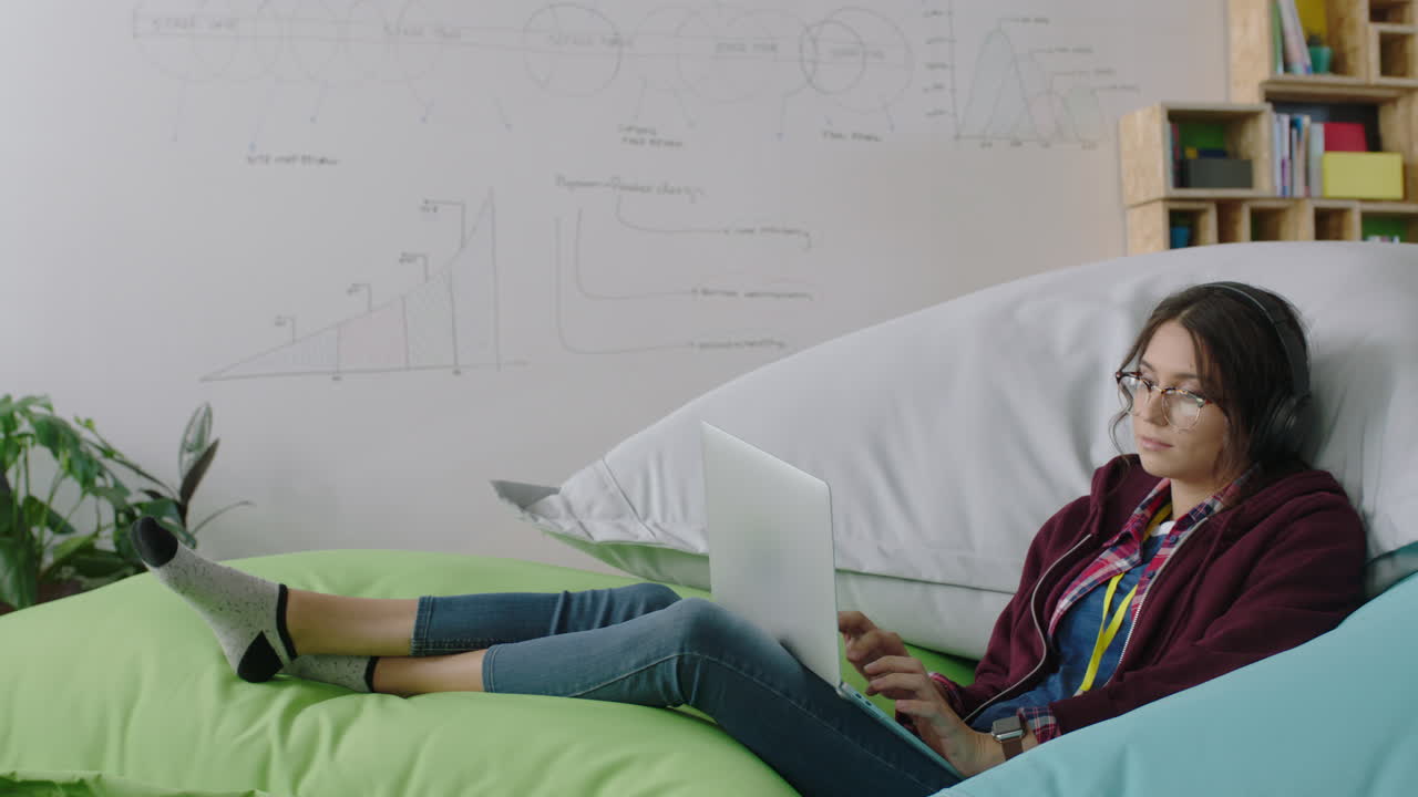 디지털 노트북 컴퓨터를 사용하는 행복한 여성 학생 온라인 소셜 미디어 메시지를 검색하고 창의적인 프로젝트를 브레인스토밍하고 다채로운 베개에 휴식을 취하고 현대적인 사무실에서 음악을 듣고 있습니다.