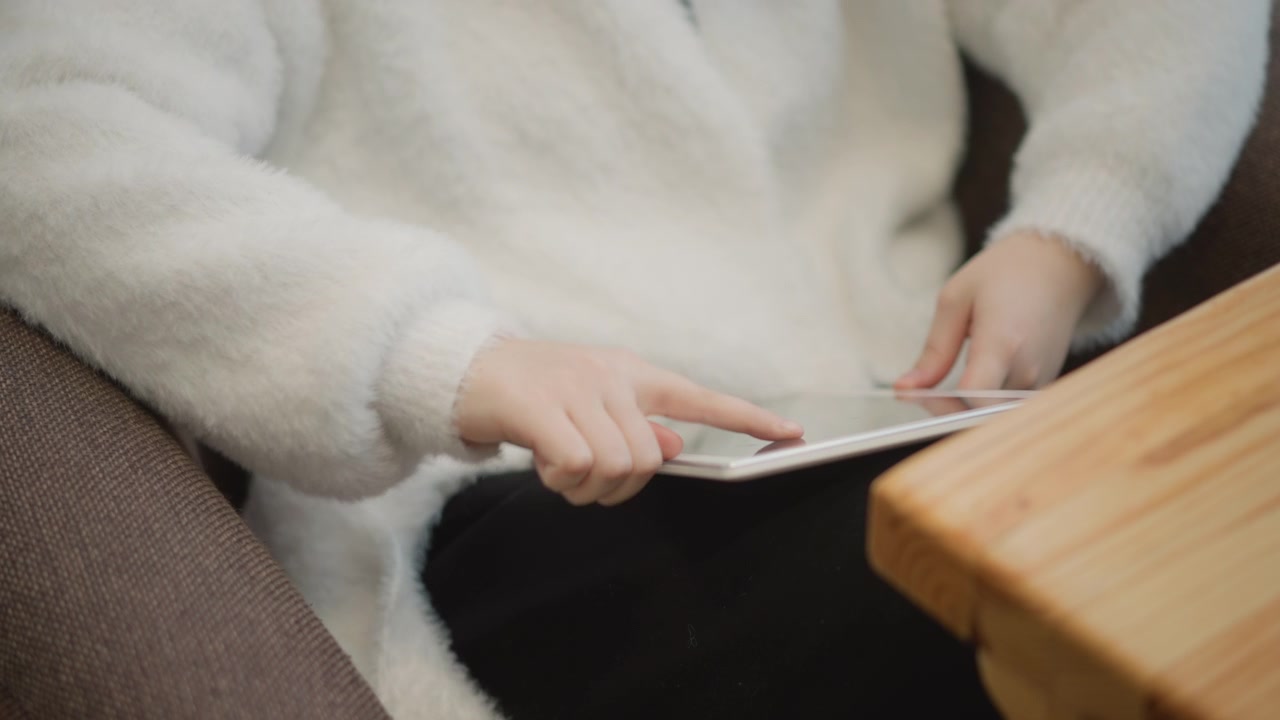Oblique top-down view of cropped torso and hands using tablet while seated indoors, capturing fingertip tapping, scrolling, and relaxed focused interaction with glowing screen, with hand, tablet
