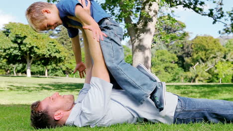 a father lifts his son above him as he lies on the grass before returning him to the ground