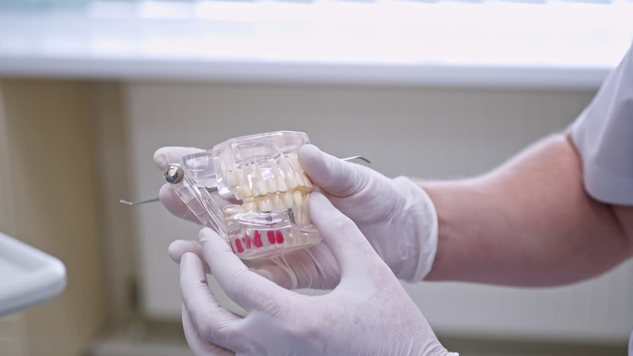 Doctor shows on a plastic jaw sample or model different methods of teeth treatment. Modern dental clinic background. Health concept. White medical gloves on male doctor's hands.