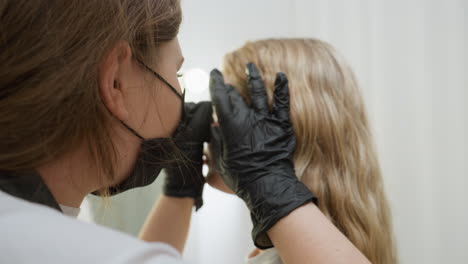 beautician in black gloves and face mask gently picks client eyebrow as blonde-haired client sits calmly in beauty salon with ring light and white curtains softly lighting the background scene