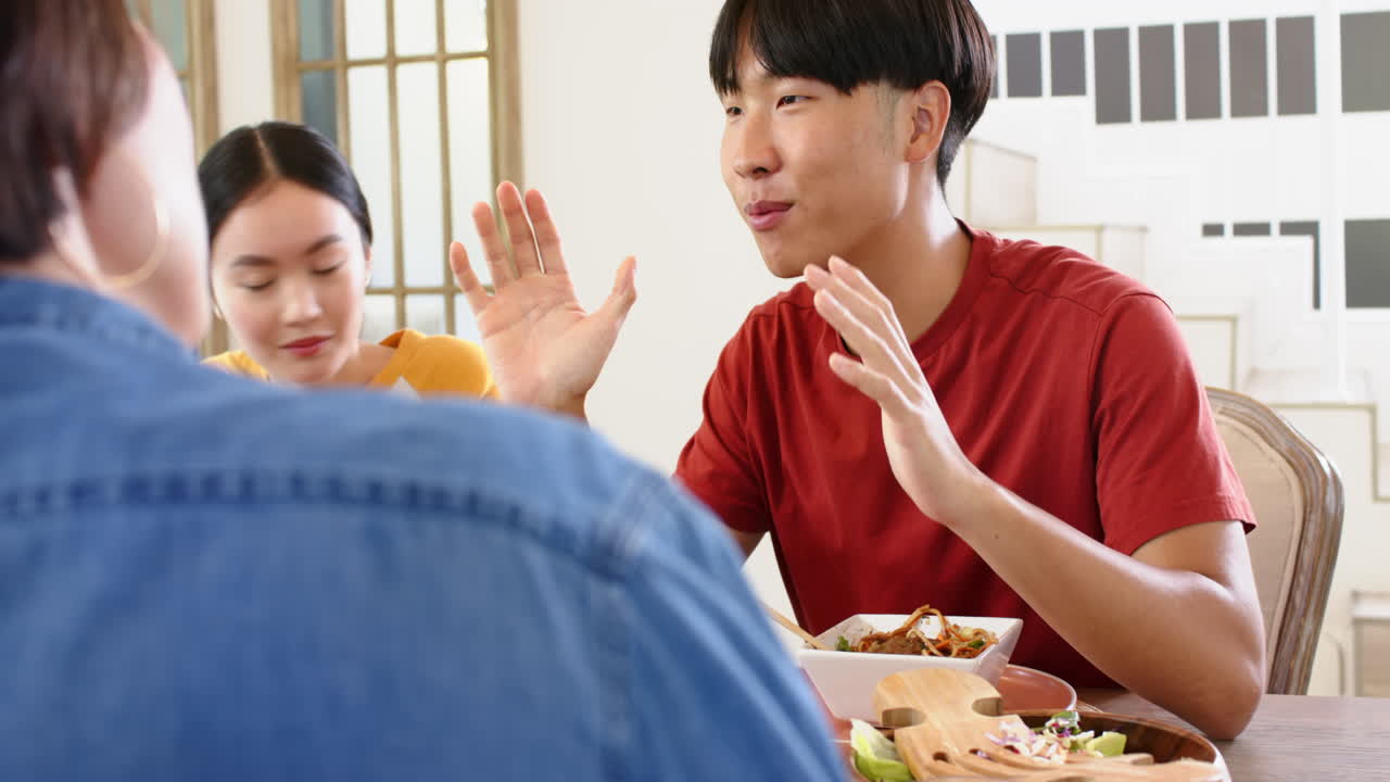 Eating together, young Asian man in red shirt talking with family at dining table