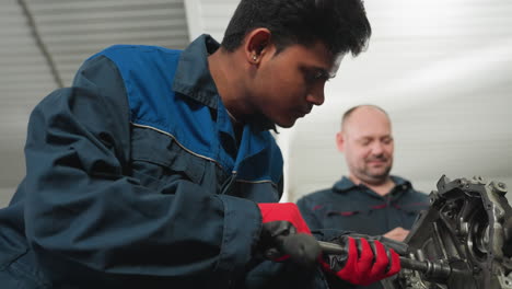 primer plano de un mecánico con pendiente magnético y guantes rojos soltando una nuez en un motor, observado atentamente por su colega en el fondo de un taller ocupado
