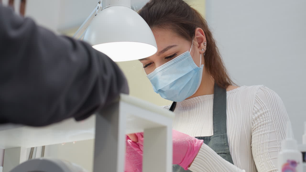 Nail technician wearing pink gloves and blue face mask attentively filing customer's nails under salon lamp, demonstrating care during manicure session in clean professional beauty salon environment