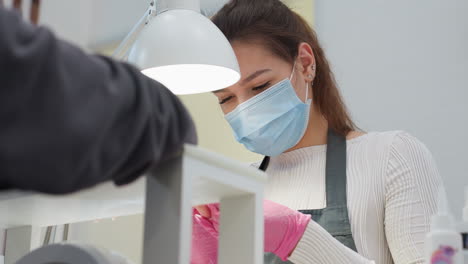nail technician wearing pink gloves and blue face mask attentively filing customer's nails under salon lamp, demonstrating care during manicure session in clean professional beauty salon environment