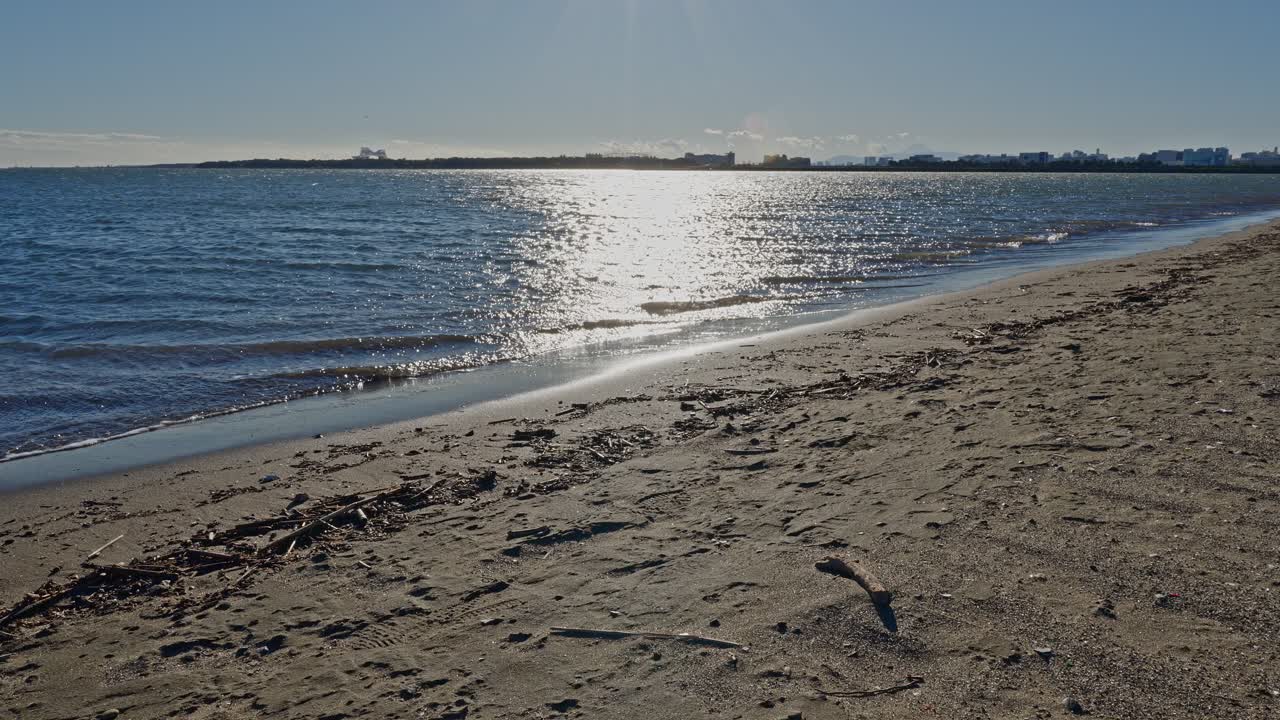 Bright, sunny view across a sandy beach and shimmering water with sun glare, showing a flat, low-rise coastline in the distance