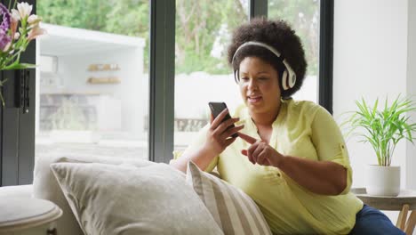 video of happy plus size african american woman with headphones sitting on sofa with smartphone