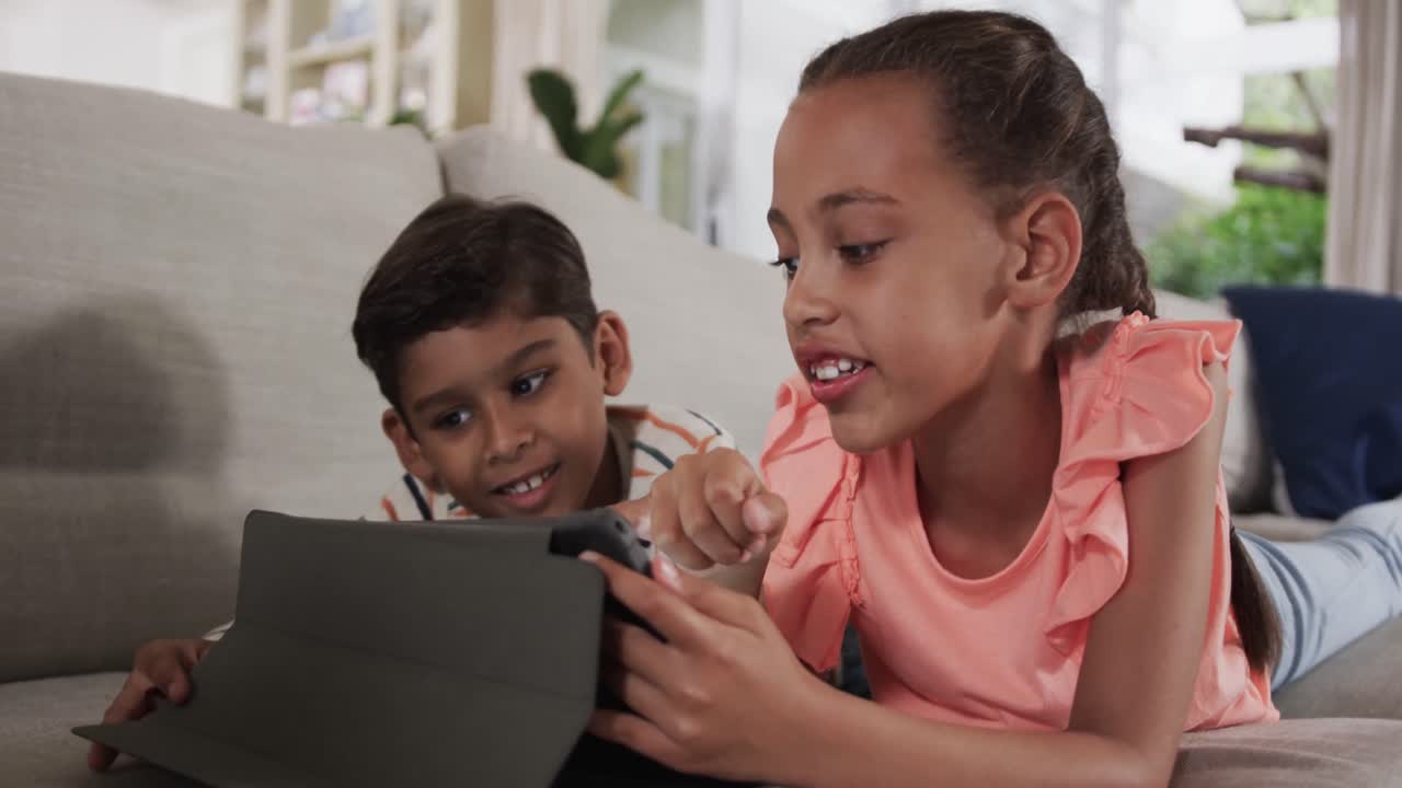 feliz hermano y hermana biracial acostados en el sofá y usando la tableta en casa, cámara lenta