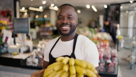 Porträt-Eines-Schwarzhäutigen-Mannes-In-Braunem-T-Shirt-Und-Schwarzer-Schürze,-Der-Mit-Gelben-Bananen-In-Den-Händen-In-Einem-Supermarkt-Posiert