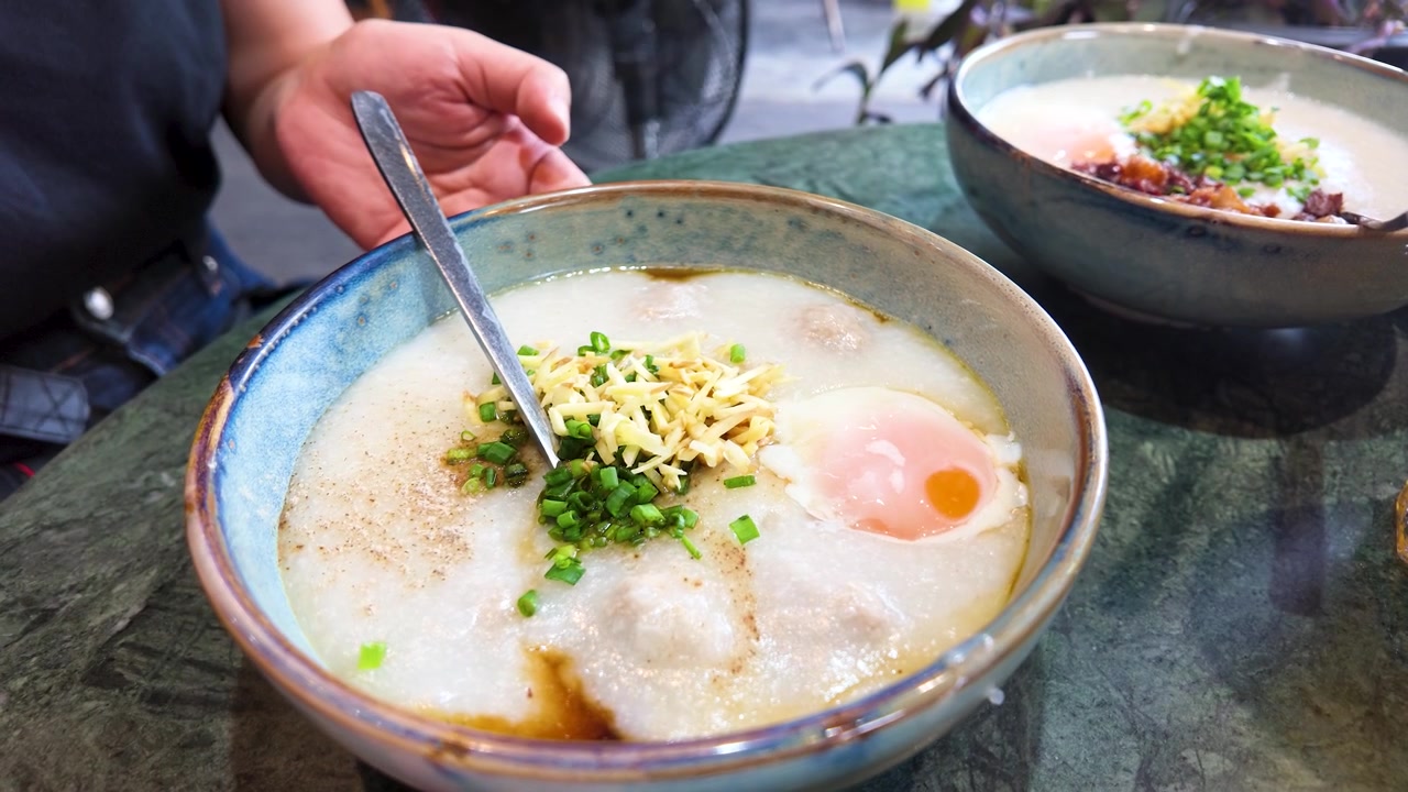 Foto close-up semangkuk bubur gurih untuk sarapan dengan jahe, daun bawang, dan telur di meja jalanan