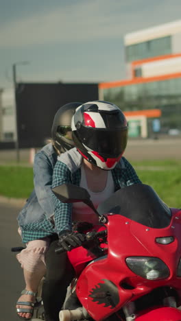 dos mujeres montando una bicicleta eléctrica roja, con la camisa del jinete revoloteando en el viento mientras su amiga se aferra por detrás, con una vista borrosa de coches, edificios y una persona caminando por una acera
