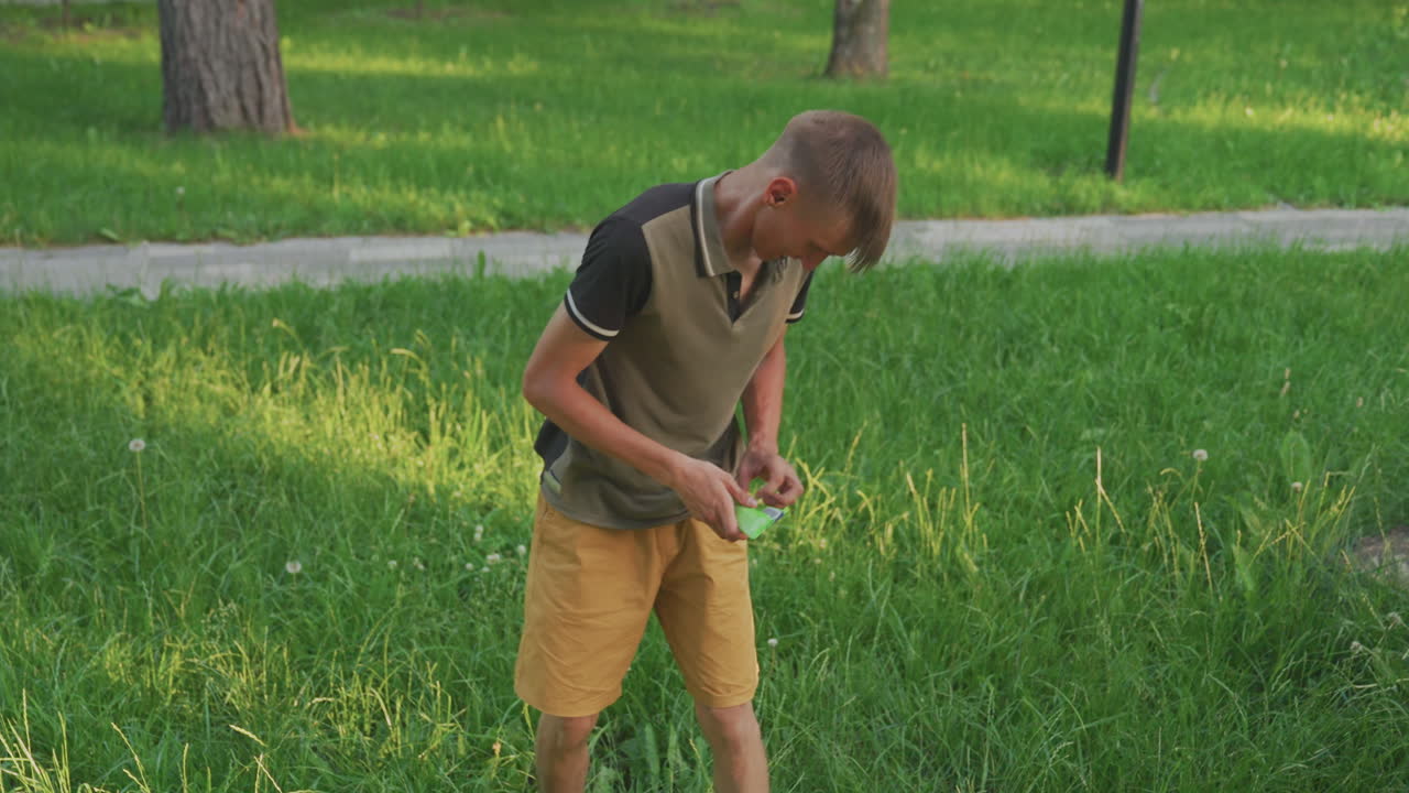 Young Man Bends To Collect Small Objects From Grassy Lawn And Rises Holding Items, Casual Polo And Shorts Visible, Park Path In Background And Warm Afternoon Light Framing The Simple Action